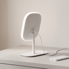Rear view of the GlowVanity mirror on a white desk with its USB charging cable running to a phone