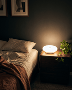 Halo donut lamp glowing warm on a bedside table next to a made bed and a plant at night