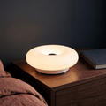 Halo Bauhaus donut lamp glowing warm on a wooden nightstand beside a bed and stacked books
