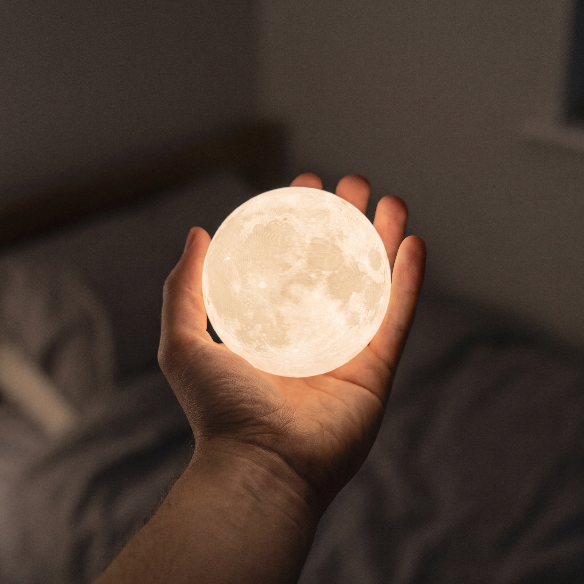 A hand holding the glowing Luna moon lamp in a dark bedroom, showing its textured crater surface