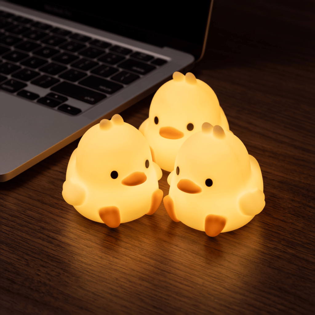 Three Peep duck and chick night lights glowing warm yellow on a wooden desk beside a laptop