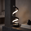 Spira black LED spiral table lamp glowing warm on a dark marble ledge beside a vase and books